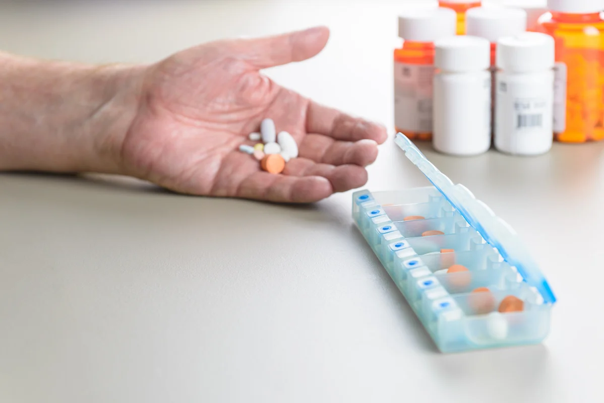 Hand holding pills beside a pill organizer and prescription bottles on a table.